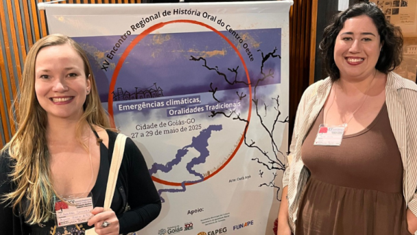 Fotografia de duas mulheres em pé, lado a lado, com os rostos desfocados, posicionadas em frente a um banner vertical. O banner anuncia o XVI Encontro Regional de História Oral do Centro-Oeste, com o tema “Emergências climáticas, Oralidades Tradicionais”, realizado na Cidade de Goiás (GO), entre os dias 27 e 29 de maio de 2025. A arte do evento é assinada por Carla Ayê. Na parte inferior do banner, constam os logotipos das instituições apoiadoras: Goiás 200 anos, FAPEG e FUNAPE.