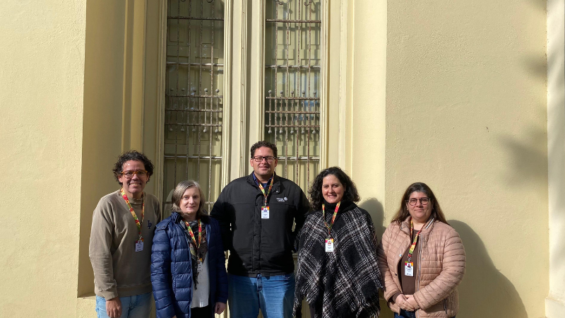 Cinco pessoas estão em pé, lado a lado, em frente a uma parede de cor clara com uma janela alta e gradeada ao fundo. Todas usam crachás pendurados no pescoço. Da esquerda para a direita: a primeira pessoa veste um suéter bege e calça jeans; a segunda, um casaco azul escuro; a terceira, uma jaqueta preta e calça jeans; a quarta, um poncho preto e branco; e a quinta, um casaco rosa claro.
