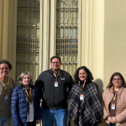 Cinco pessoas estão em pé, lado a lado, em frente a uma parede de cor clara com uma janela alta e gradeada ao fundo. Todas usam crachás pendurados no pescoço. Da esquerda para a direita: a primeira pessoa veste um suéter bege e calça jeans; a segunda, um casaco azul escuro; a terceira, uma jaqueta preta e calça jeans; a quarta, um poncho preto e branco; e a quinta, um casaco rosa claro.
