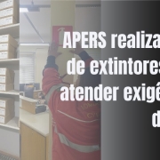 A imagem mostra um ambiente interno com foco em segurança contra incêndios. À esquerda, há um extintor de incêndio vermelho fixado na parede, com uma placa de sinalização acima dele. Ao lado, vêem-se prateleiras contendo diversas caixas de arquivo organizadas. No centro da imagem, uma pessoa usando uniforme vermelho está instalando ou inspecionando outro extintor de incêndio em uma parede. À direita, há um texto em destaque que diz: "APERS realiza troca de extintores para atender exigências do PPCI".