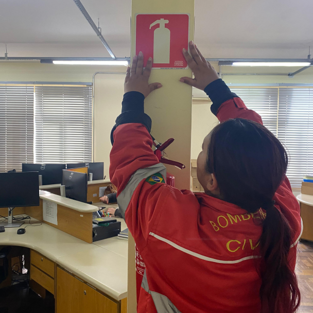 A imagem mostra o interior de um escritório. Em primeiro plano, uma pessoa usando uniforme vermelho com a inscrição "BOMBEIRO CIVIL" nas costas está fixando uma placa vermelha com o símbolo de um extintor de incêndio em uma coluna. Ao fundo, há mesas com computadores e cadeiras, e as janelas estão com as persianas fechadas.